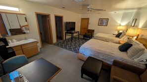 A hotel room with a bed, a desk, a chair, a small table, a sofa, a TV, and a bathroom with a mirror and sink. - Home-like Cabin Room - Room 144 (Custer)