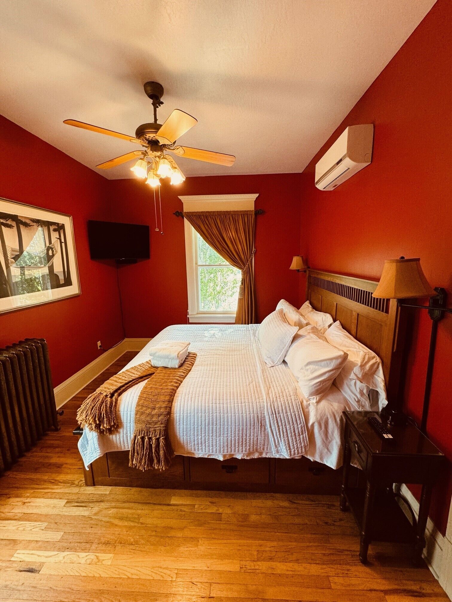 Un dormitorio con cama, ventilador de techo, ventana con cortinas, radiador, televisor y lámpara.