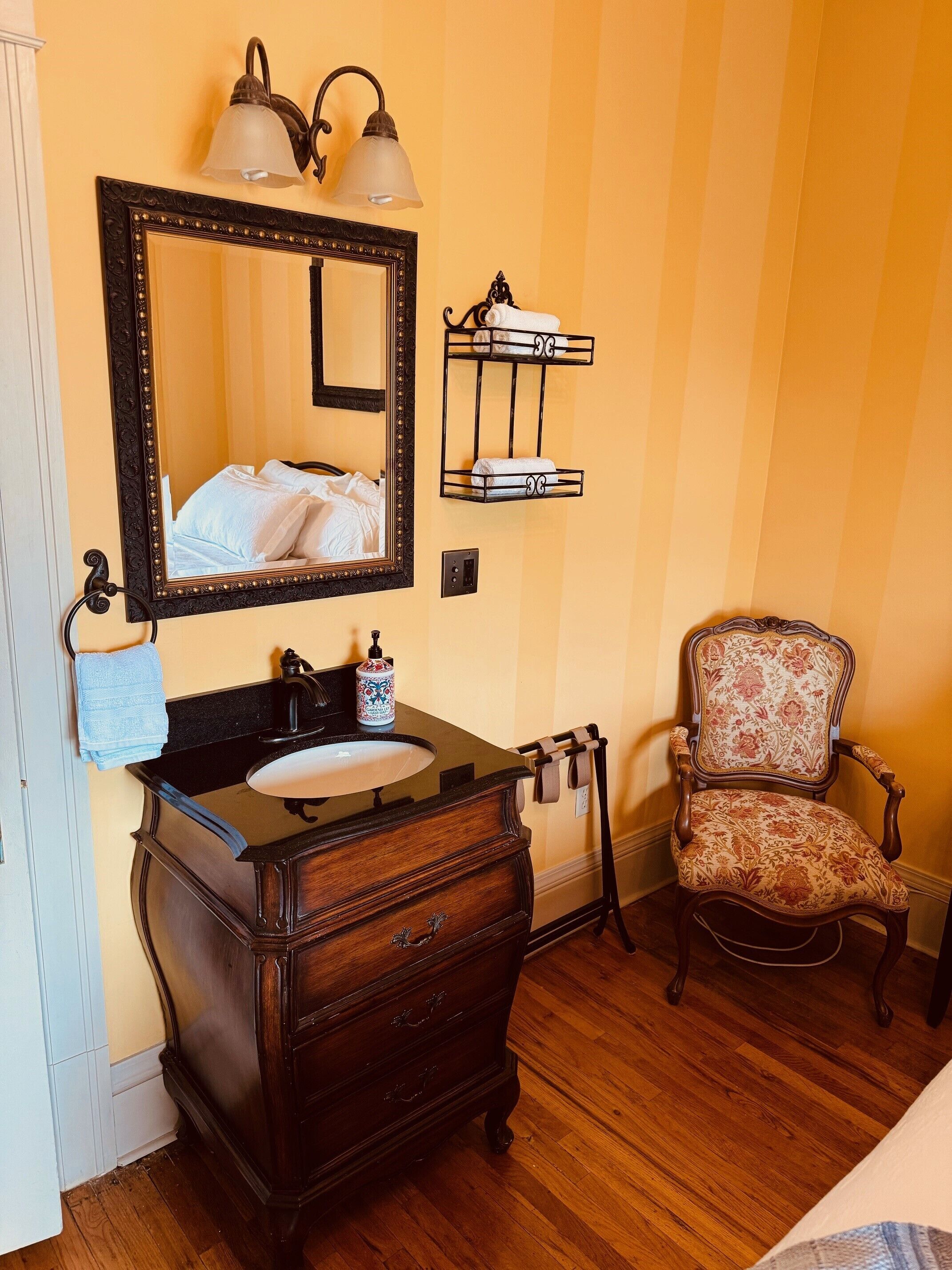 A bathroom with a wooden vanity, a large mirror, a towel rack, and a floral chair.
