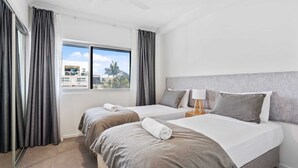A hotel room with two beds, a large window with curtains, a ceiling fan, and a view of buildings and palm trees outside. - Sunrise Cove (Mermaid Beach)