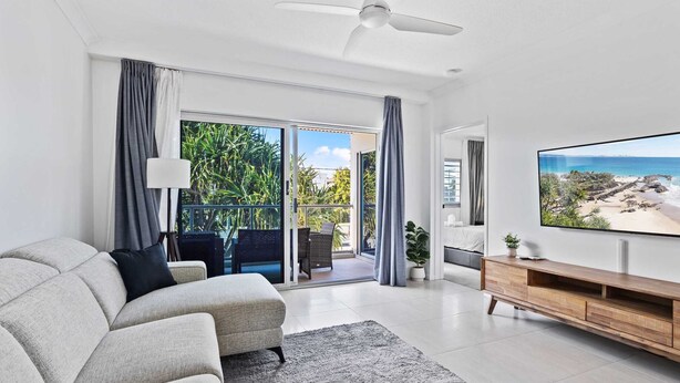 A modern living room with a sofa, a flat-screen TV, and a balcony with a view of the ocean. - Sunrise Cove (Mermaid Beach)