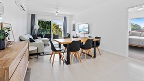 A modern living room with a wooden dining table, black chairs, a grey sofa, and a TV on the wall. - Sunrise Cove (Mermaid Beach)