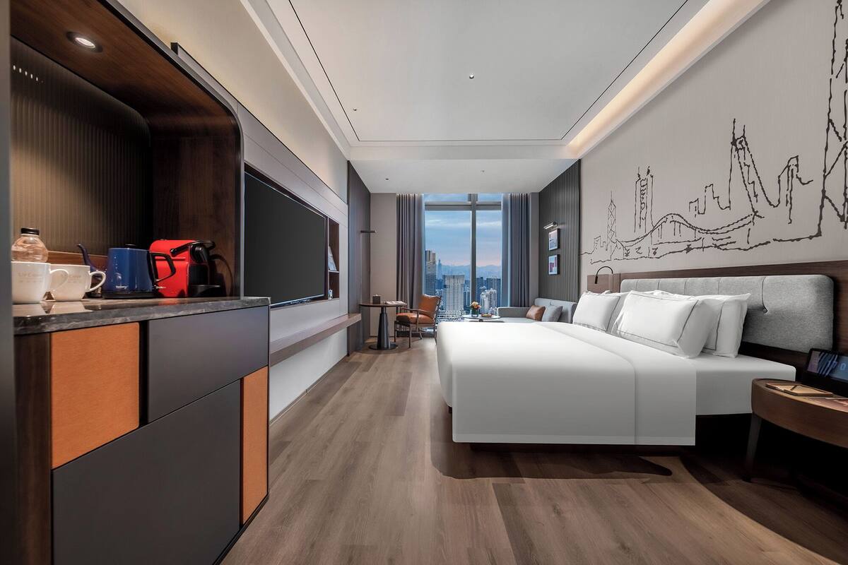 Une chambre d’hôtel moderne dotée d’un grand lit, d’un bureau avec un téléviseur à écran plat, d’une vue sur la ville grâce à de grandes fenêtres et d’un mur orné d’une fresque représentant un paysage urbain.