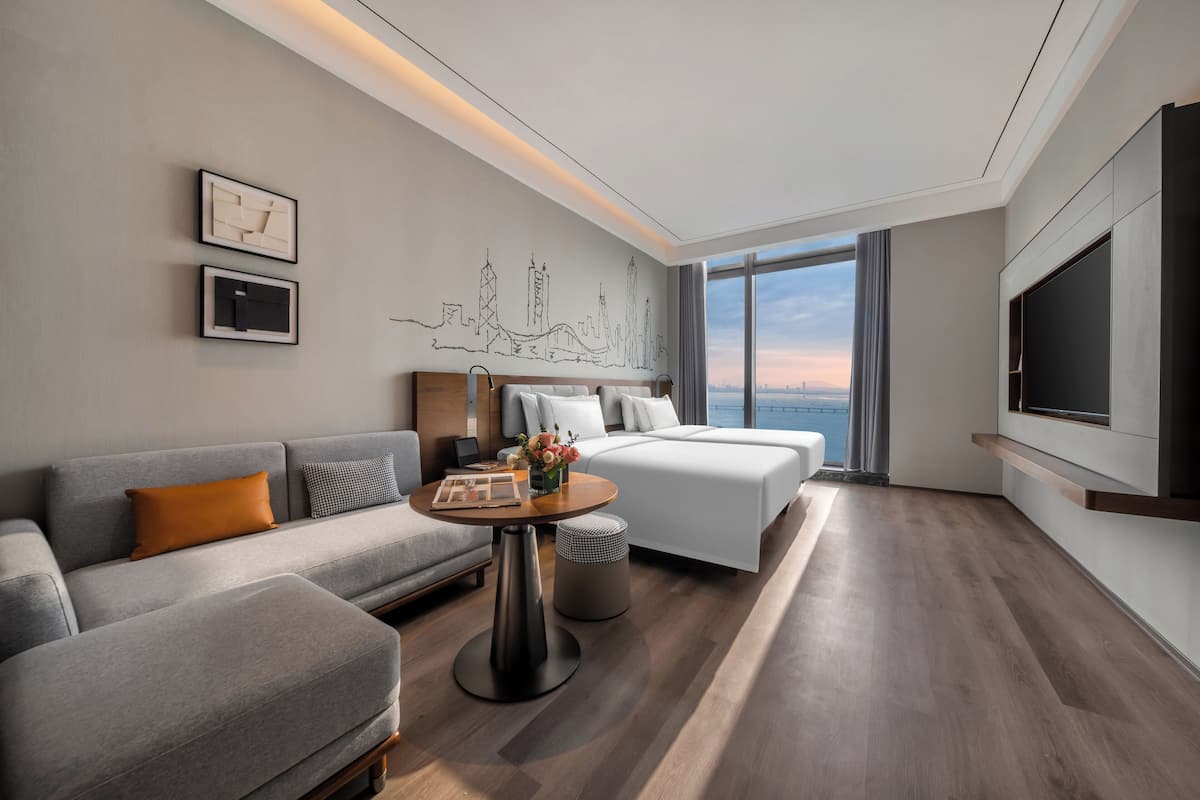 Une chambre d’hôtel moderne, équipée d’un canapé, d’un lit, d’une petite table sur laquelle est posé un vase de fleurs et d’un grand téléviseur.