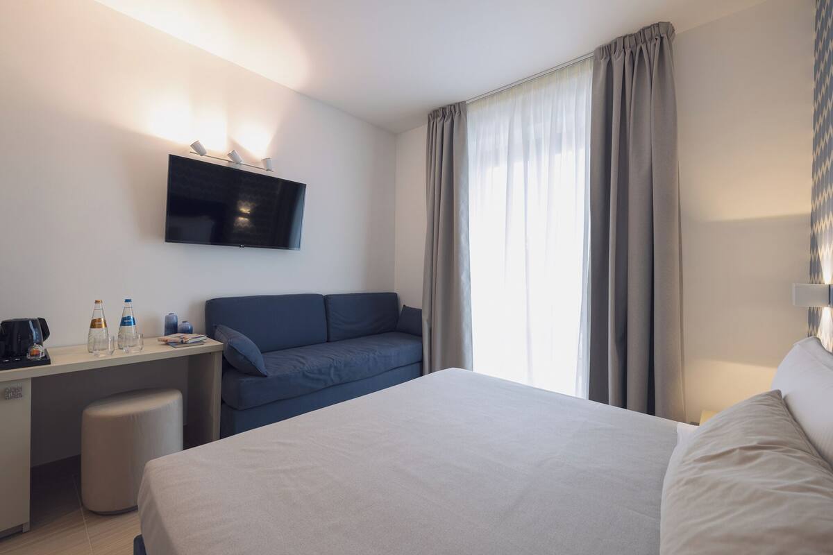 A hotel room with a bed, a desk with a flat-screen TV, a sofa, and a window with curtains.