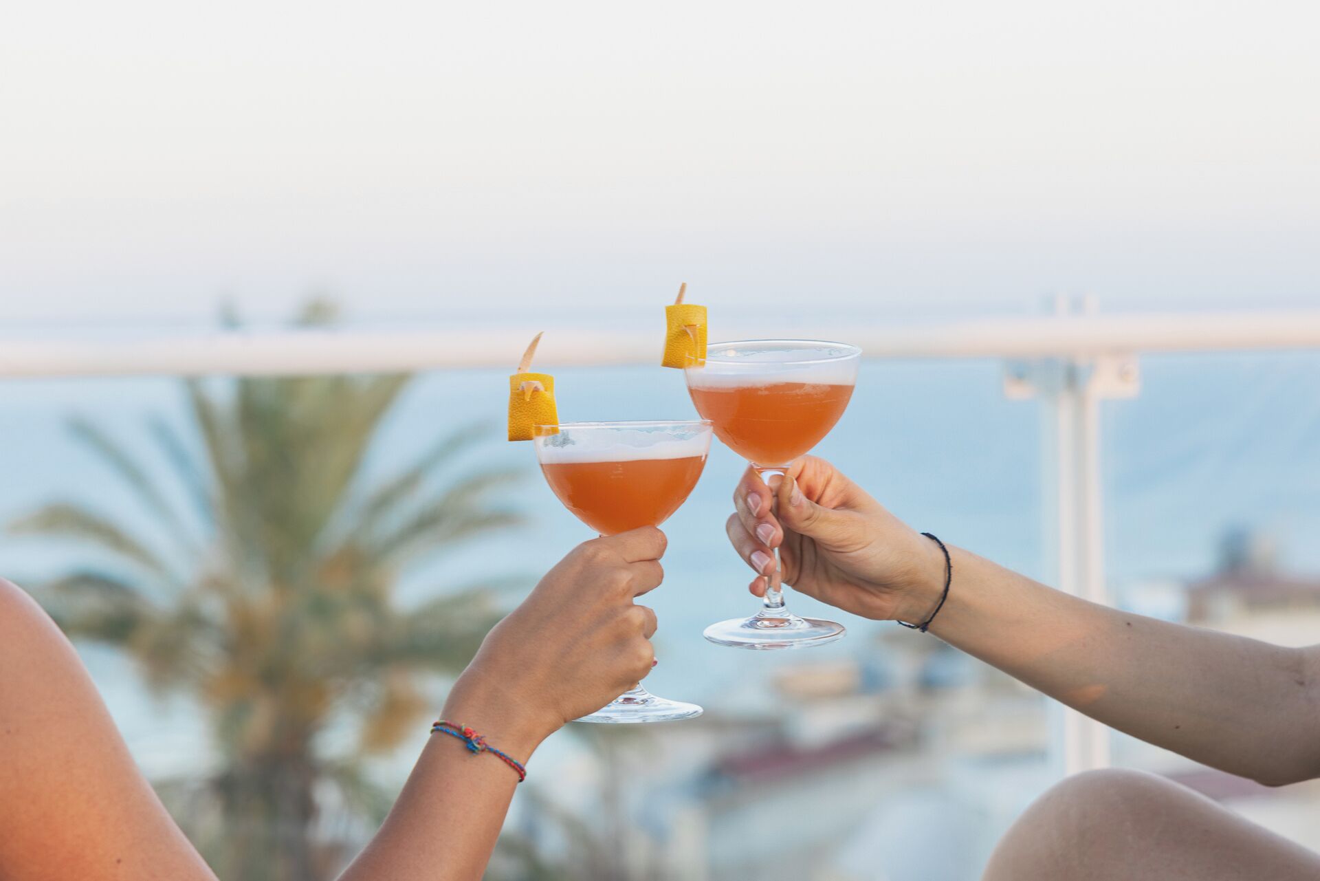 Twee personen proosten met cocktails op een balkon met uitzicht op een wateroppervlak.