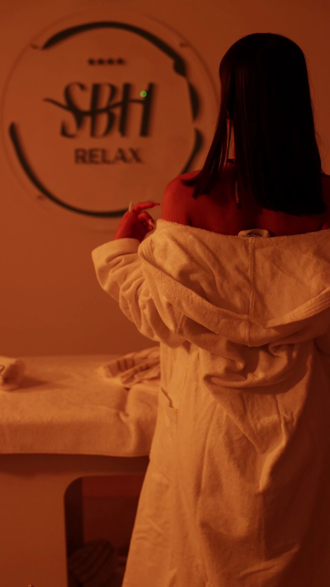 A person in a white robe standing in a room with a circular sign that reads "SBB RELAX".