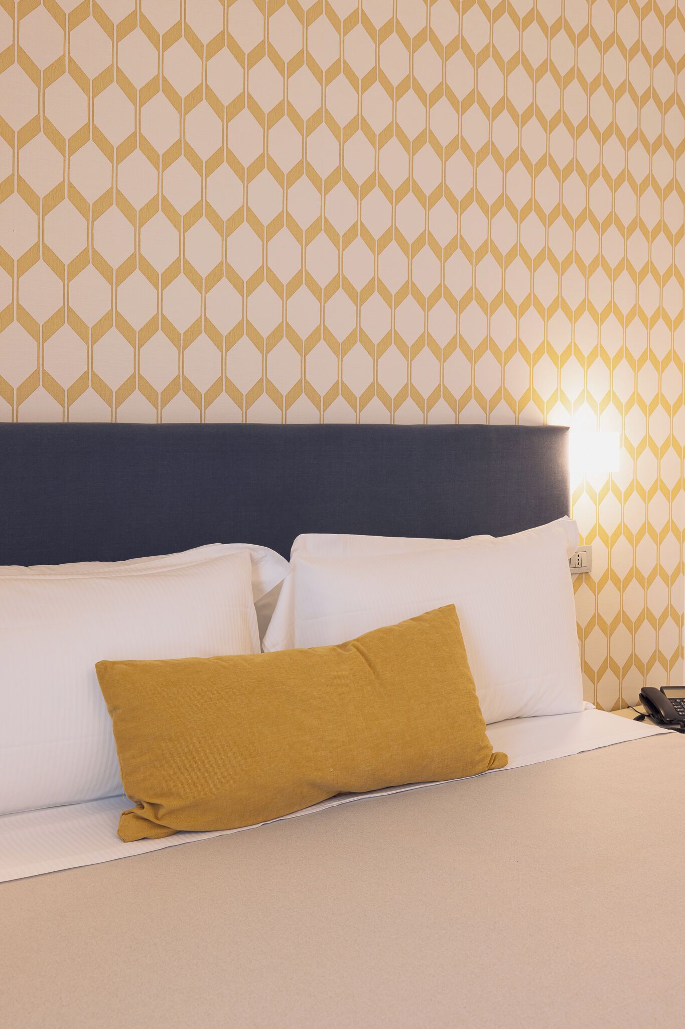 Una habitación de hotel con papel tapiz estampado, cabecera oscura y cama con almohadas blancas y mostaza.