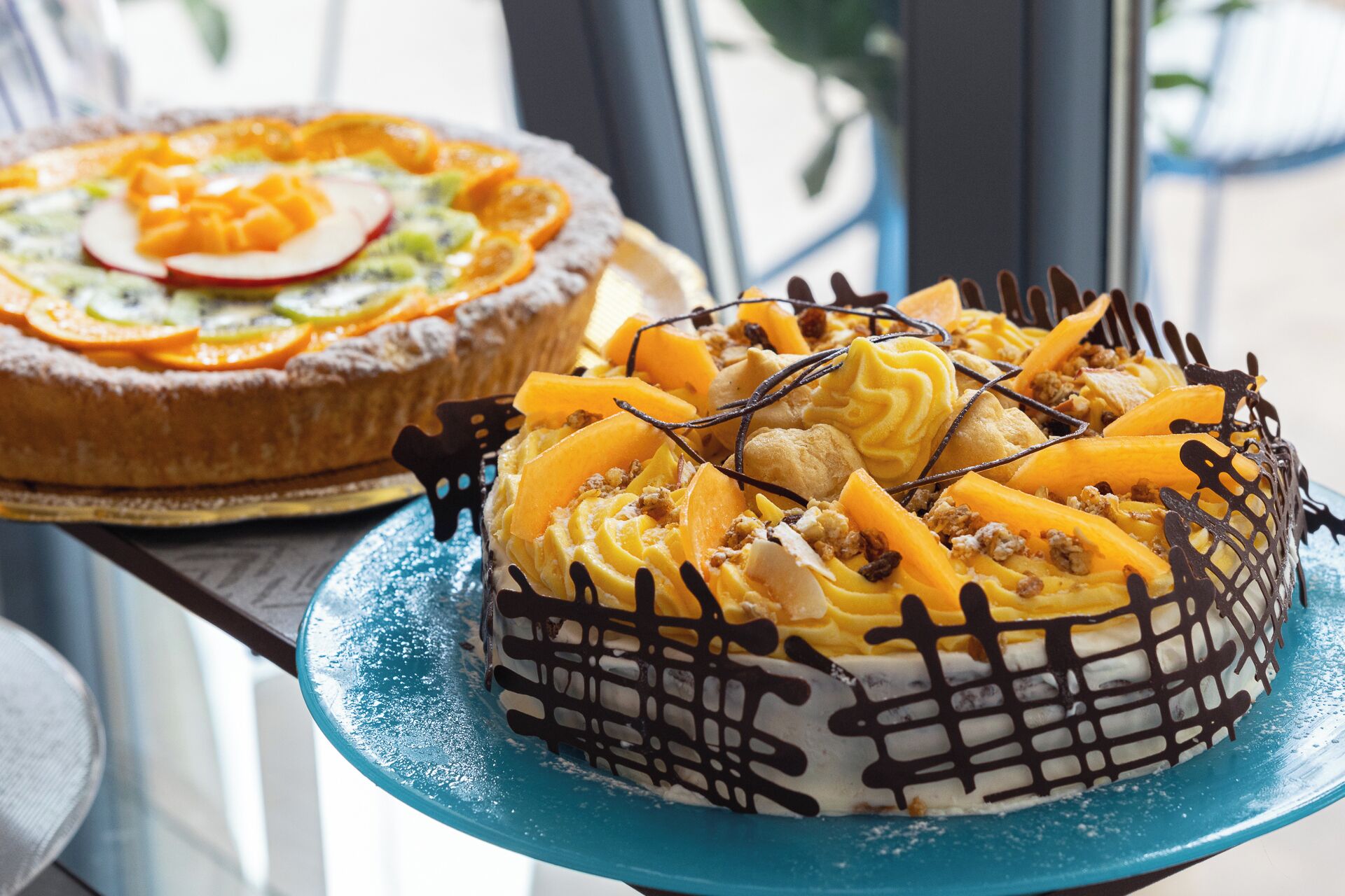 A dessert display with a cake topped with fruit and chocolate drizzle, and another cake in the background.