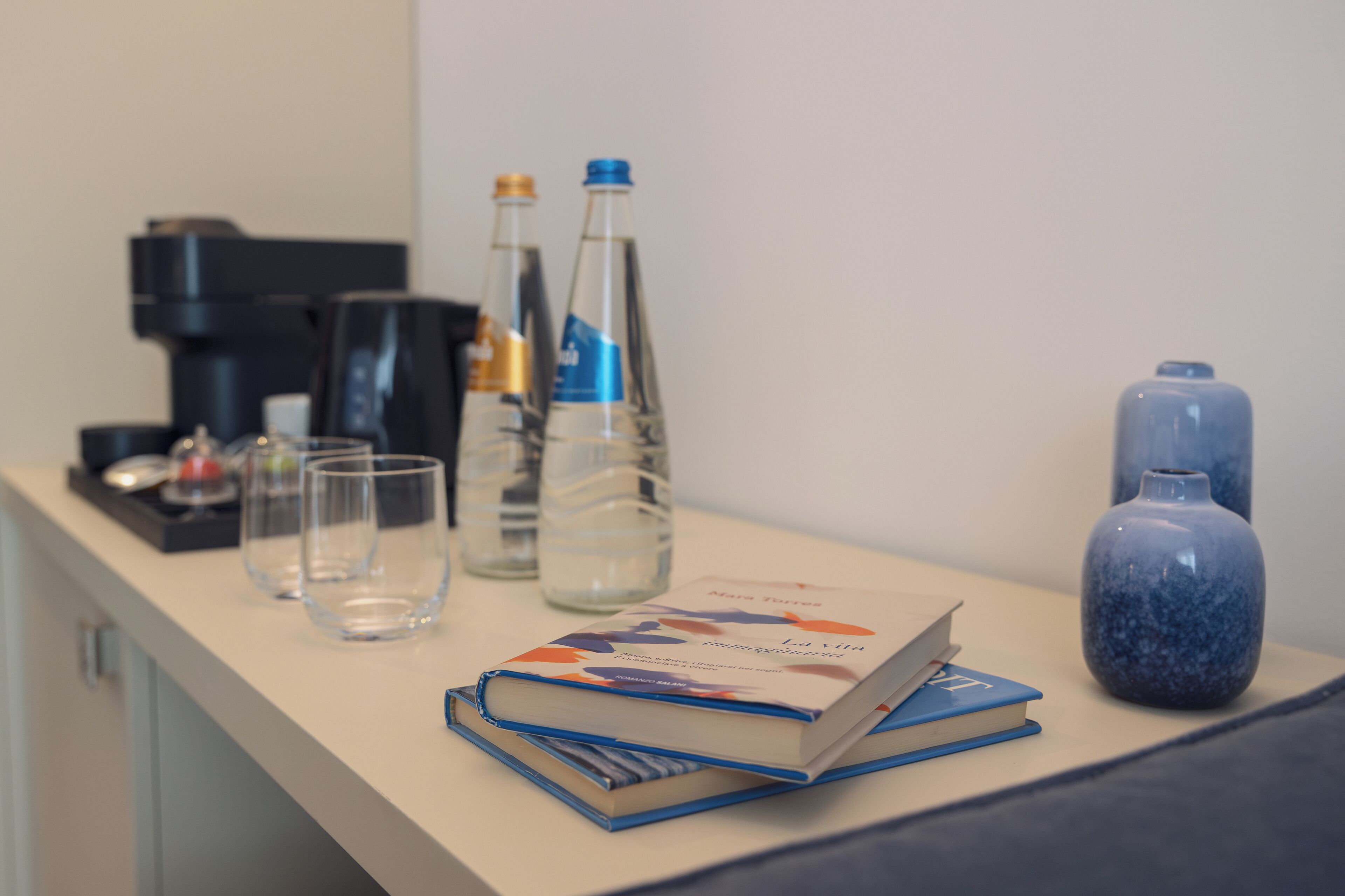A hotel room with a coffee maker, two water bottles, a vase, and a stack of books on a table.