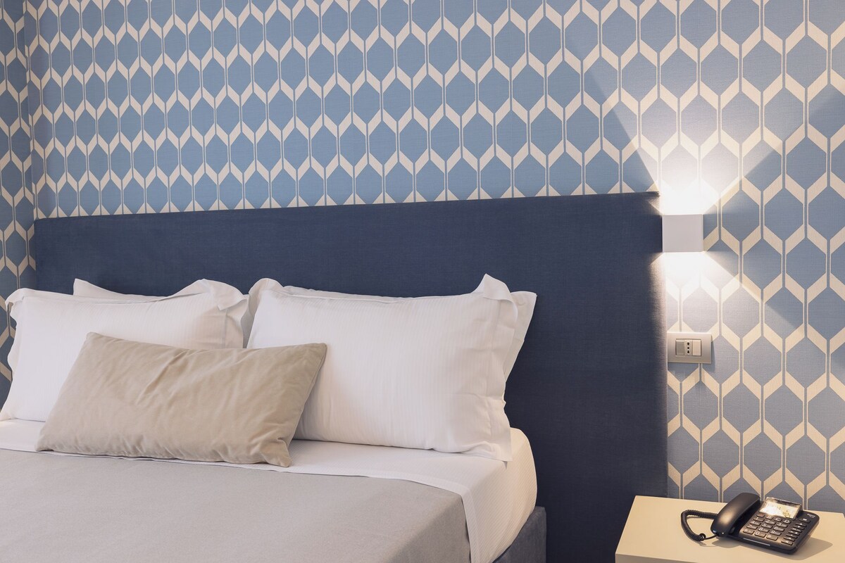 A neatly made bed with white linens and a beige pillow, a bedside telephone, and a wall-mounted light fixture.