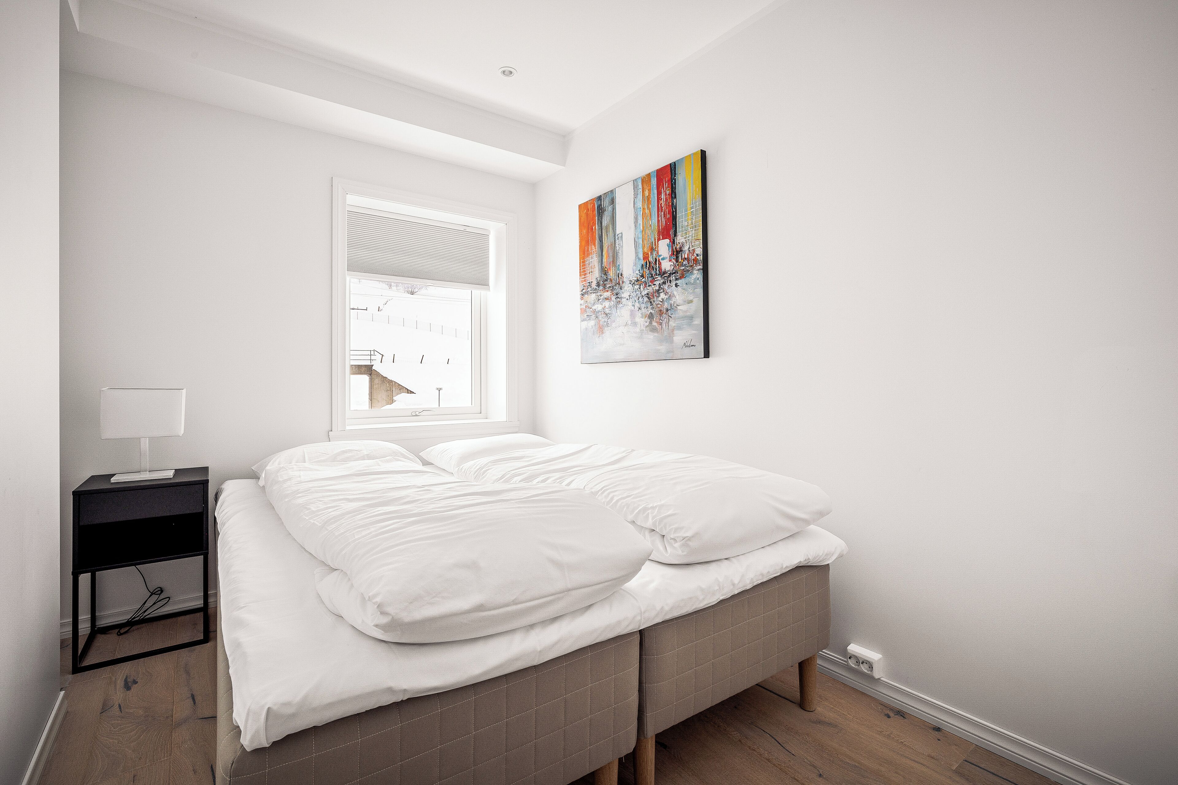 A bedroom with a bed, a nightstand, a window, and a painting on the wall.