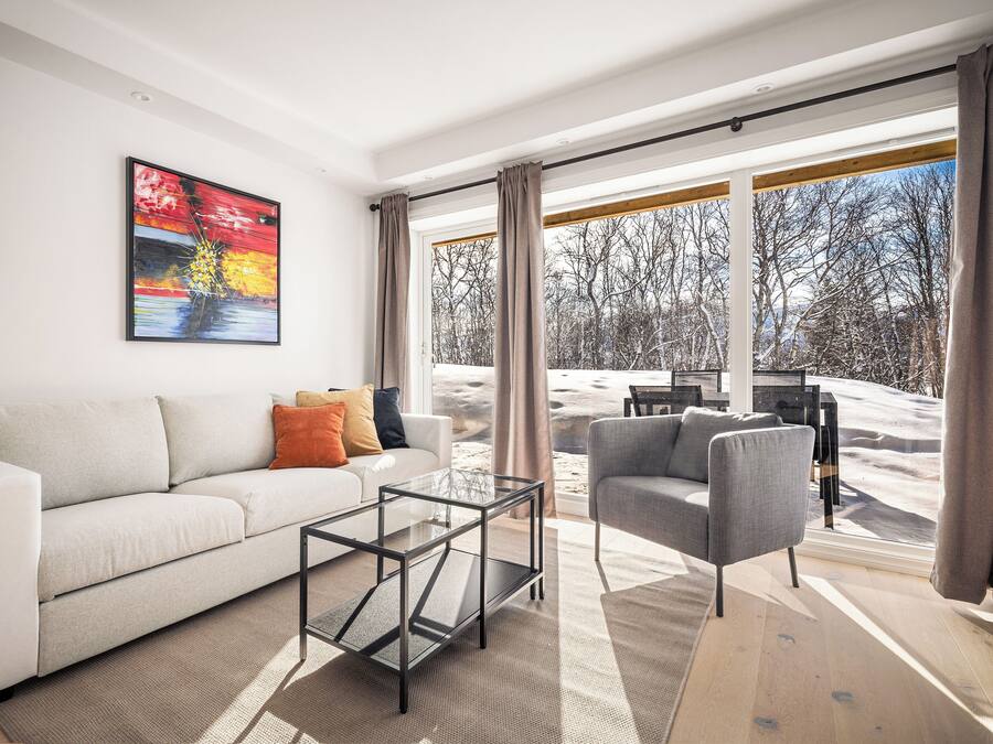 Un salón moderno con un sofá, una mesa de centro de vidrio y un sillón gris. Hay un cuadro en la pared y una ventana grande con cortinas.
