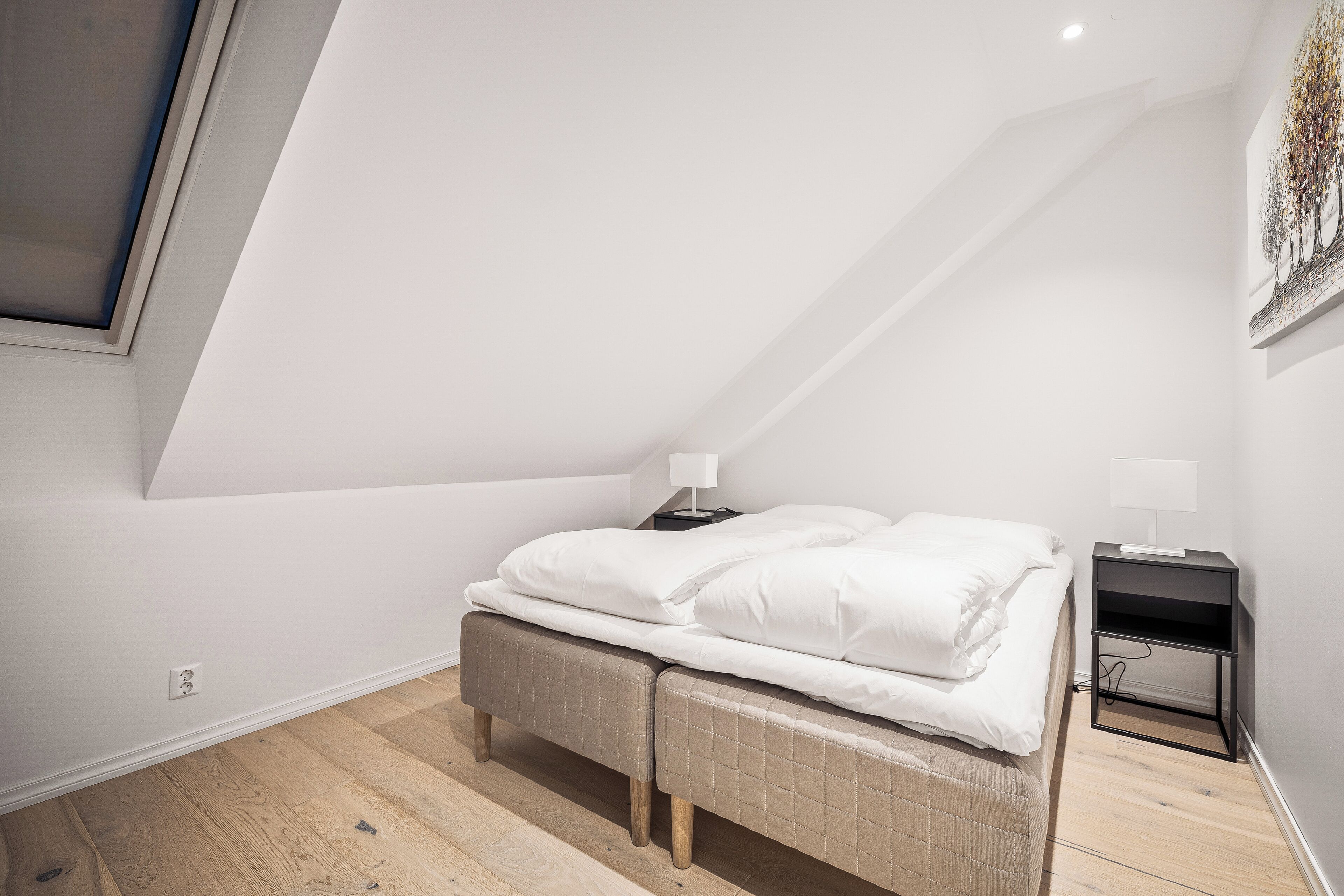 A bedroom with a sloped ceiling, a bed with white bedding, a nightstand with a lamp, and a wall-mounted artwork.