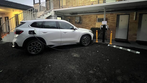 Un SUV blanco estacionado en una estación de carga frente a un edificio de fachada de ladrillo y una escalera que sube a un nivel superior.