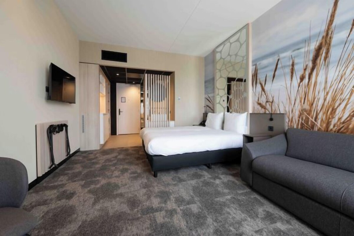 A hotel room with a bed, a sofa, a TV, and a painting of tall grass.
