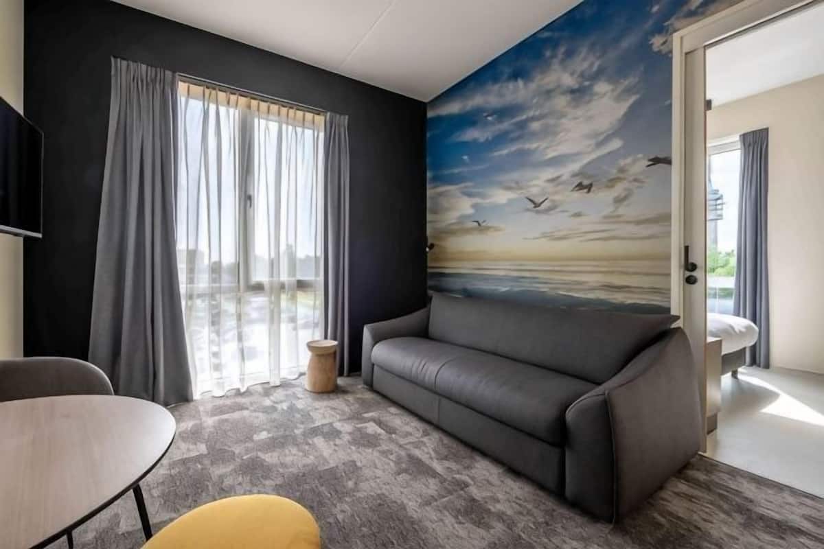 Ein Zimmer mit einem grauen Sofa, einem kleinen runden Tisch und einem Wandgemälde, das eine Strandansicht zeigt.