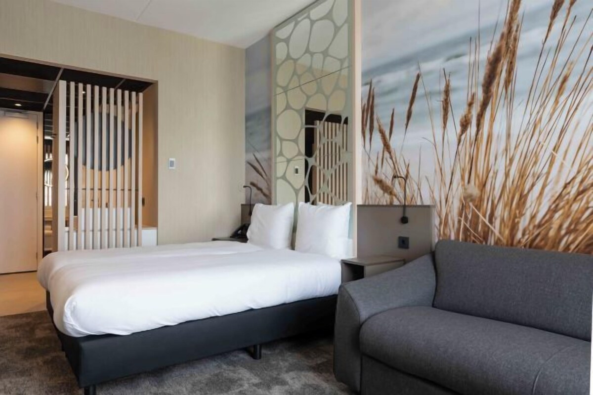 Una habitación de hotel moderna con cama, un sofá gris, una mesita de noche y una pared con diseño decorativo.