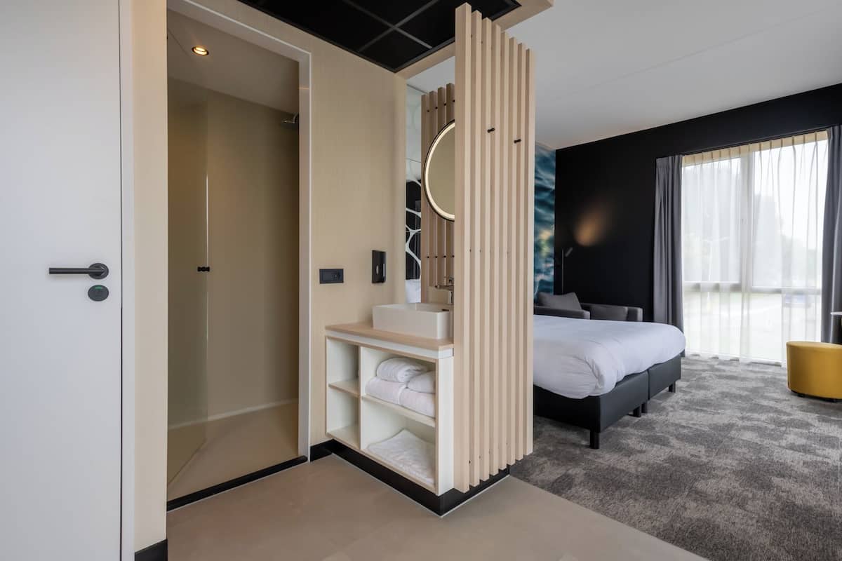 Ein modernes Hotelzimmer mit Bett, Badezimmer mit Dusche, Schminktisch mit Spiegel und Blick ins Freie.