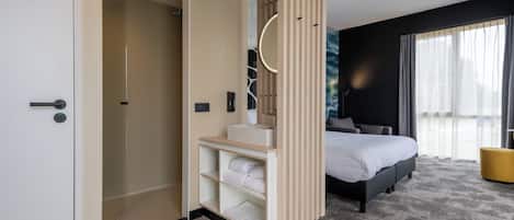 A modern hotel room with a bed, a bathroom with a shower, a vanity with a mirror, and a view of the outdoors.