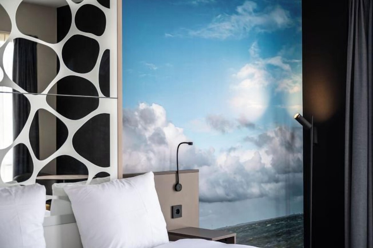 A modern hotel room with a bed, white pillows, a decorative wall panel, and a view of a cloudy sky.