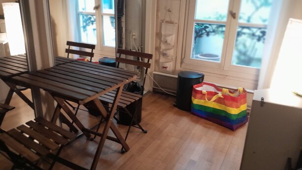 Une pièce avec une table et des chaises en bois, une poubelle noire et un sac aux couleurs de l’arc-en-ciel posé sur le sol.