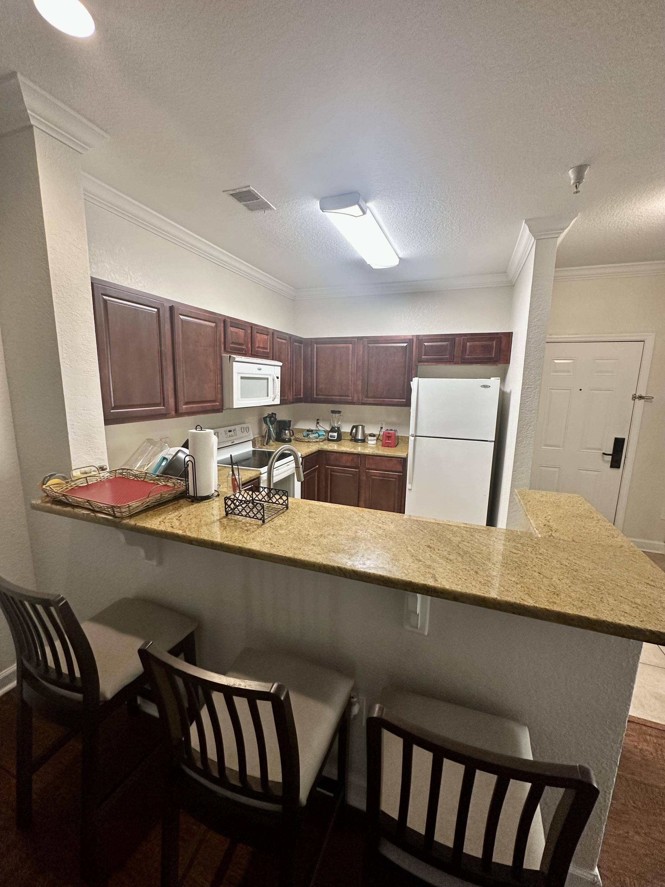 A kitchen with dark wooden cabinets, a white refrigerator, a microwave, and a granite countertop. There are two bar stools and a small island with a sink.