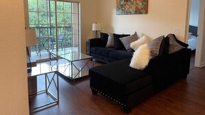 Ein Wohnzimmer mit einem dunkelgrauen Sofa, einem Glastisch, einer Lampe und einem Gemälde an der Wand.