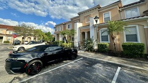 A parking lot with a black sports car, a row of beige townhouses with white trim, and a stop sign. - Grand Panoramic 3BR Townhouse Near Disney (Kissimmee)