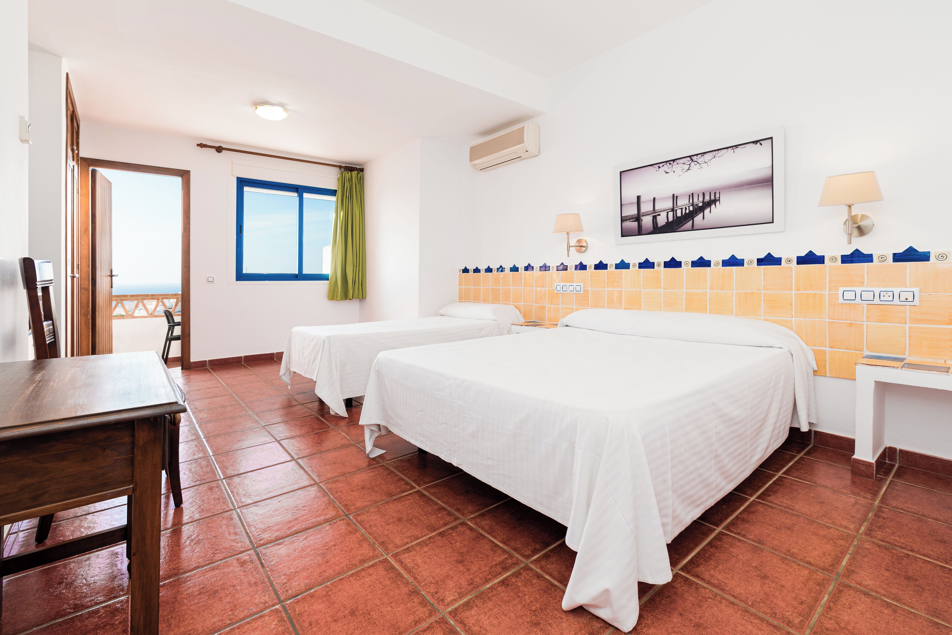 A hotel room with two beds, a wooden table, a window with curtains, a wall-mounted air conditioner, a wall-mounted lamp, and a tiled backsplash.