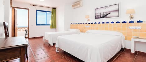A hotel room with two beds, a wooden table, a window with curtains, a wall-mounted air conditioner, a wall-mounted lamp, and a tiled backsplash.