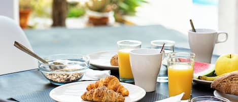 Una mesa de desayuno con cruasanes, zumo de naranja, una taza blanca, un vaso de zumo de naranja, un bol de cereales, un plato con una rebanada de sandía y un cuenco pequeño de mermelada.