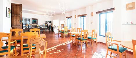 A spacious dining area with wooden tables and chairs, a kitchen in the background, and large windows allowing natural light.