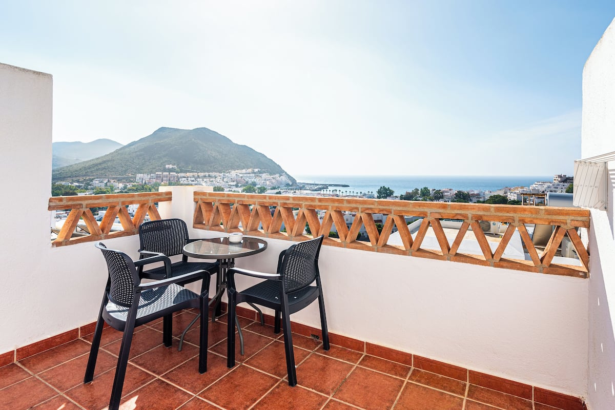 Un balcon avec une table et des chaises, offrant une vue sur la côte, avec des montagnes et des bâtiments en arrière-plan.