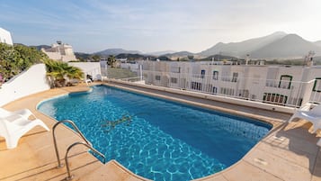 A rooftop pool with clear blue water, surrounded by white buildings and a mountainous backdrop.