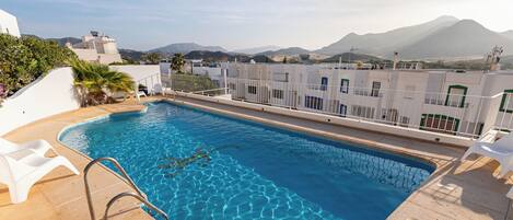 A rooftop pool with clear blue water, surrounded by white buildings and a mountainous backdrop.