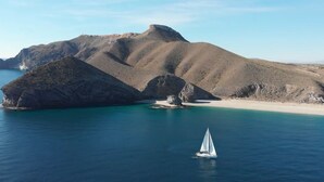 A coastal landscape with a sailboat in the water, rocky hills in the background, and a sandy beach. - El Dorado San Jose (San José)