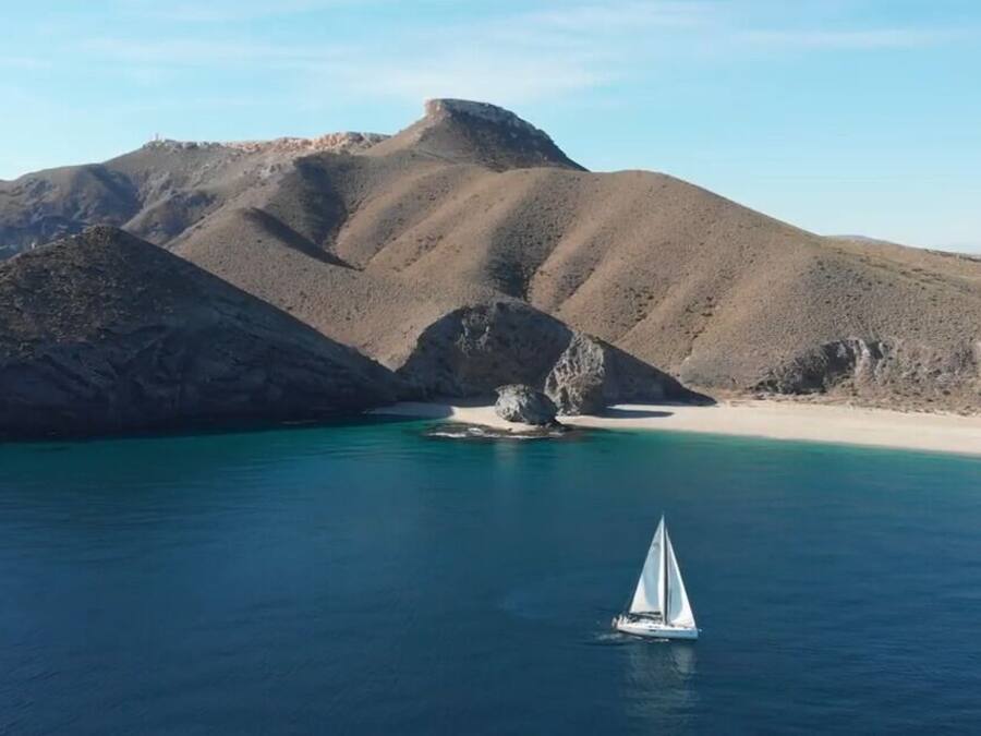 Paisagem costeira com um veleiro na água, colinas rochosas ao fundo e uma praia de areia.