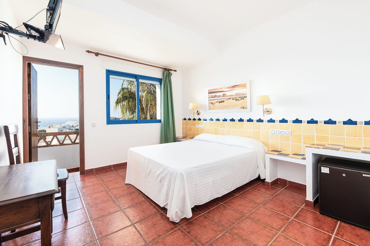 Une chambre d’hôtel comprenant un lit, un bureau, une chaise, un petit réfrigérateur, une fenêtre avec vue et un mur orné d’un tableau et de deux lampes murales.