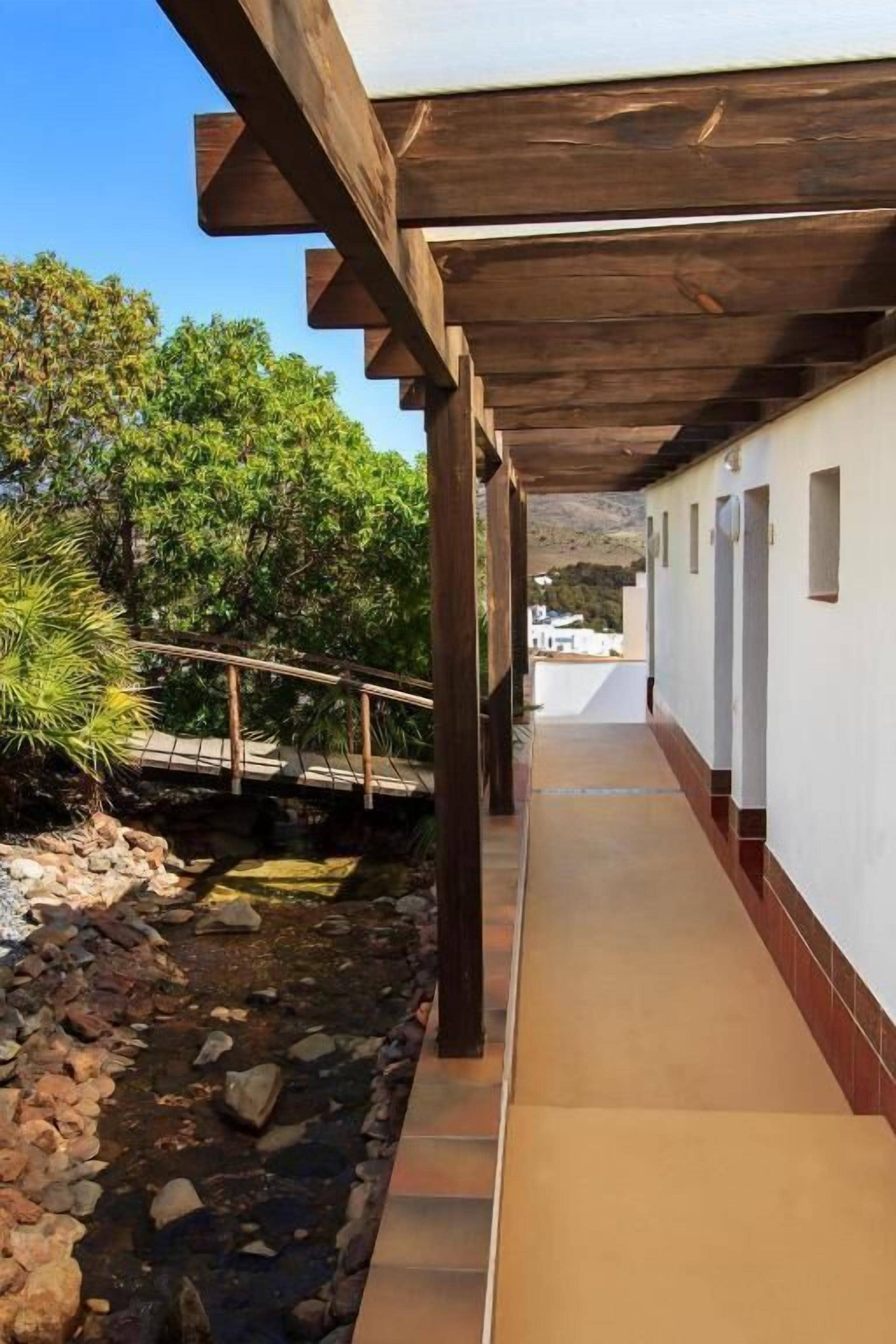 Un paseo techado con un sendero de piedra y un puente de madera sobre un arroyo, que conduce a un edificio con paredes blancas y suelo de baldosas.