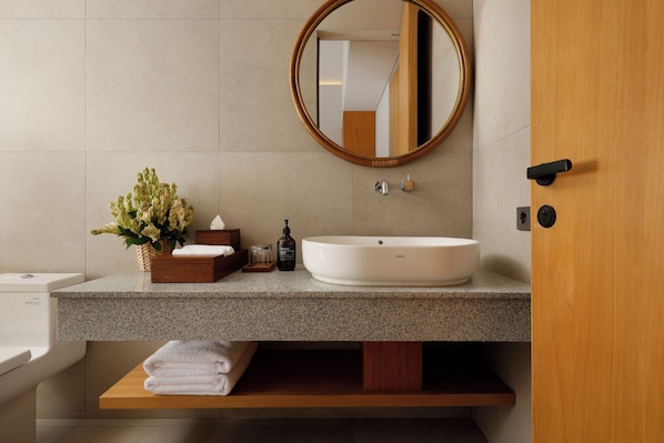 A modern bathroom with a freestanding sink, a round mirror, a wooden door, and a shelf with folded towels.