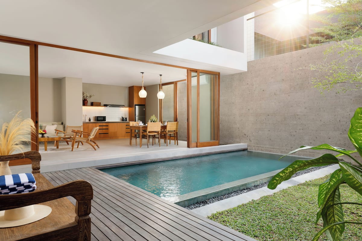 A modern outdoor pool area with a wooden deck, a dining area with chairs and a table, and a kitchen in the background.