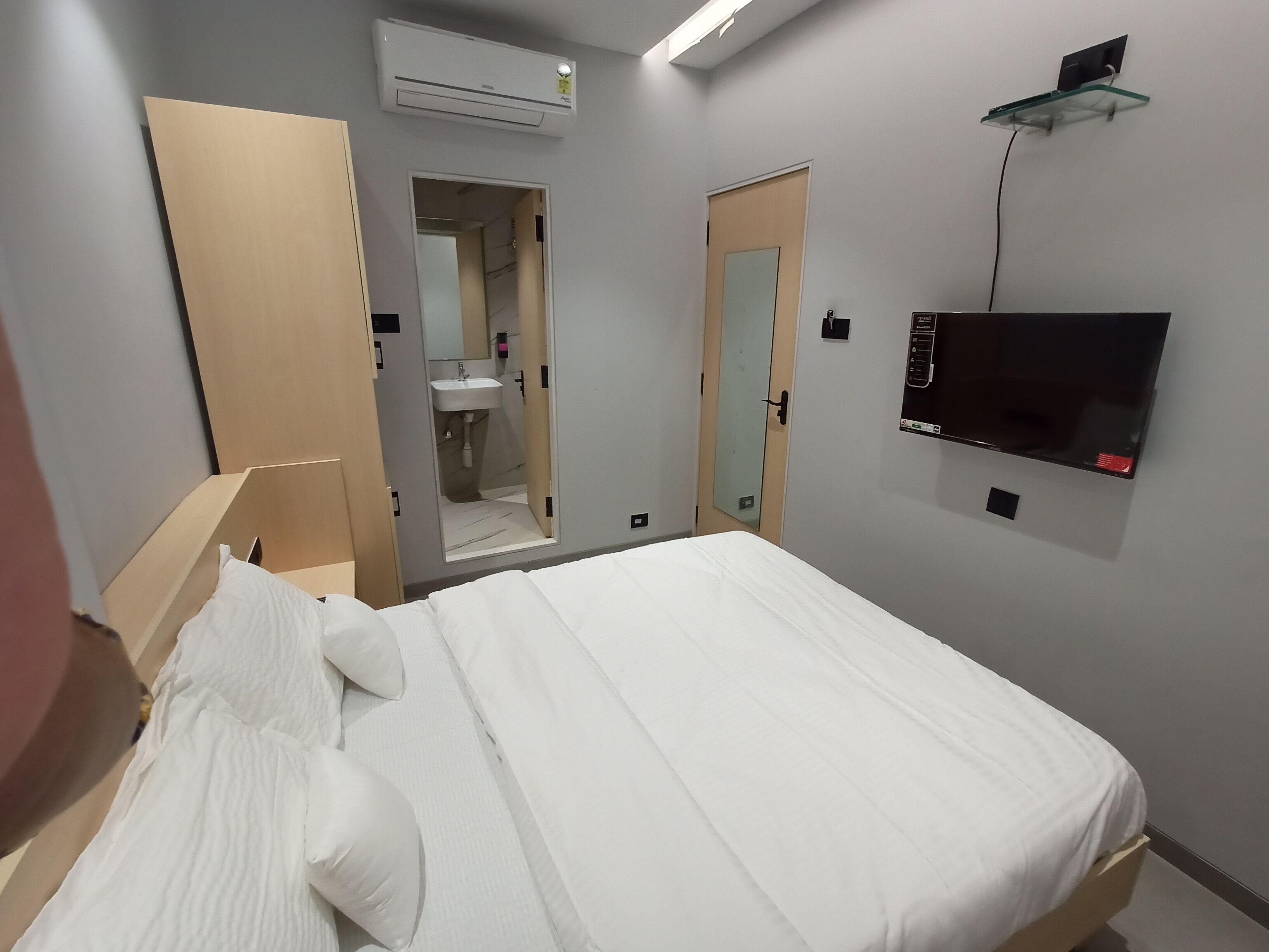 A hotel room with a bed, a TV, an air conditioning unit, a mirror, and a door leading to a bathroom.