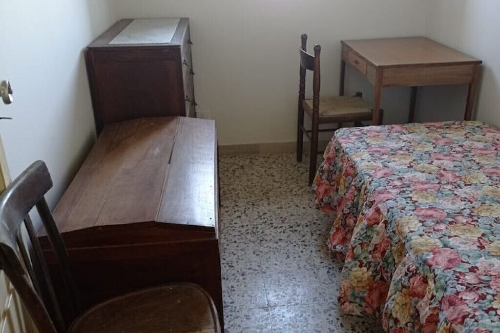 A small, sparsely furnished room with a wooden desk, a chair, a bed with a floral bedspread, and a tiled floor.