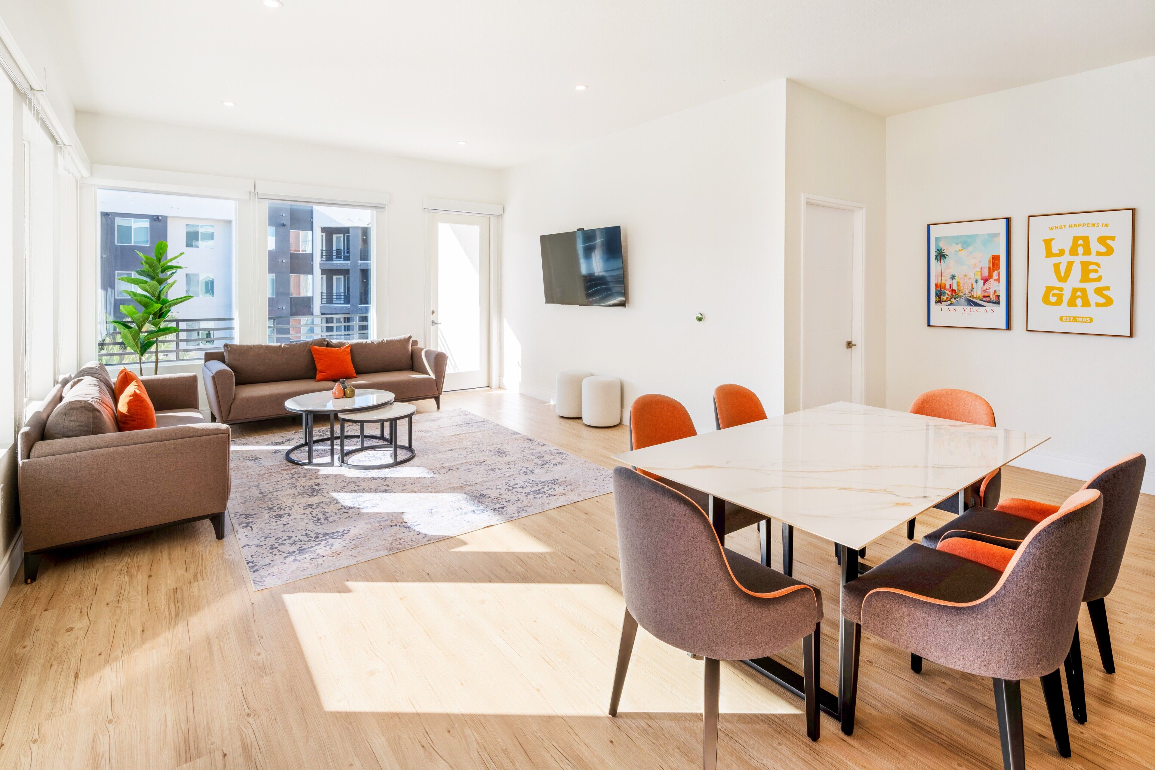 A modern living room with a sofa, coffee table, and dining table with chairs. There are large windows, a plant, and artwork on the walls.