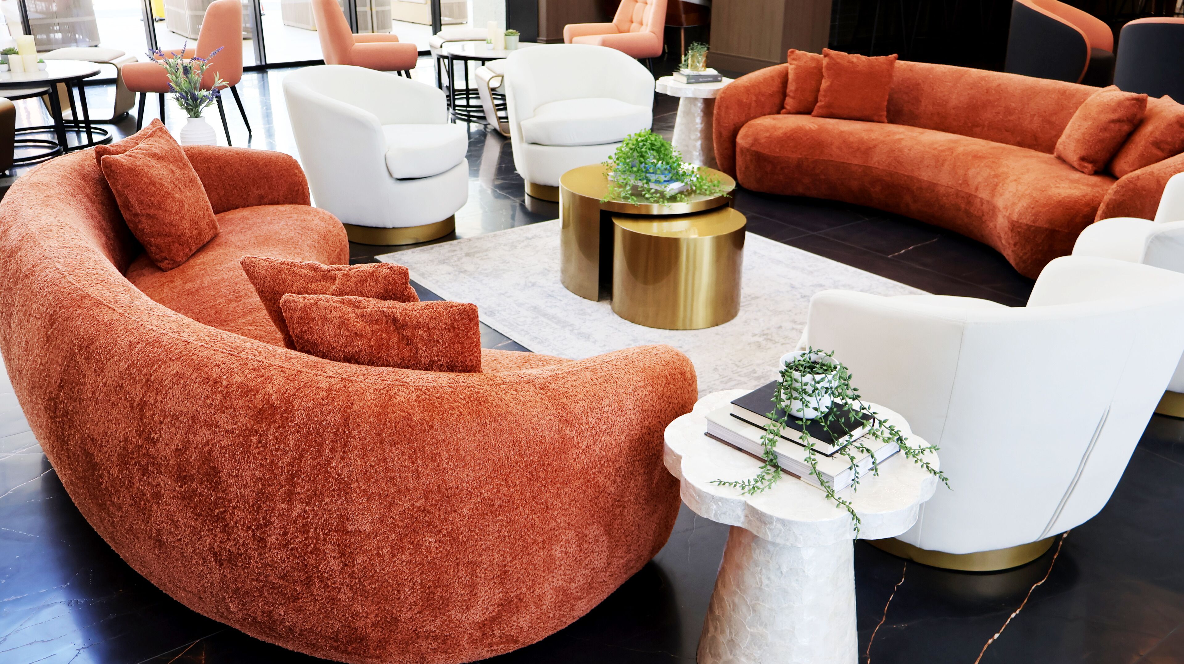 Een moderne woonkamer met een grote oranje bank, witte fauteuils en een gouden salontafel met een plant.