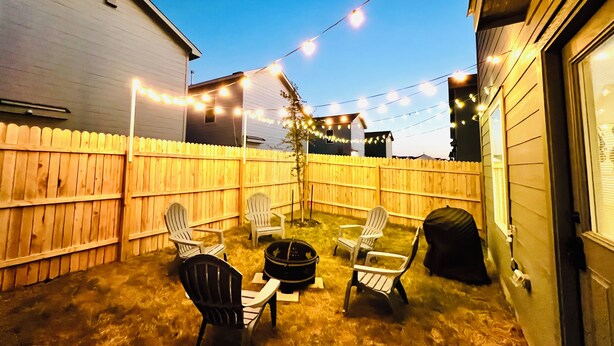 Ein Garten mit Holzzaun, Lichterketten und Gartenmöbeln, darunter Stühle und ein Tisch.