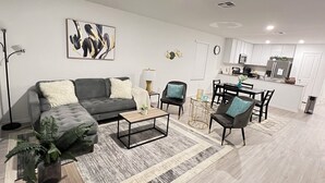 Ein modernes Wohnzimmer mit grauem Sofa, Couchtisch und Sesseln, ein Essbereich mit Tisch und Stühlen sowie eine Küche mit weißen Schränken und Geräten.