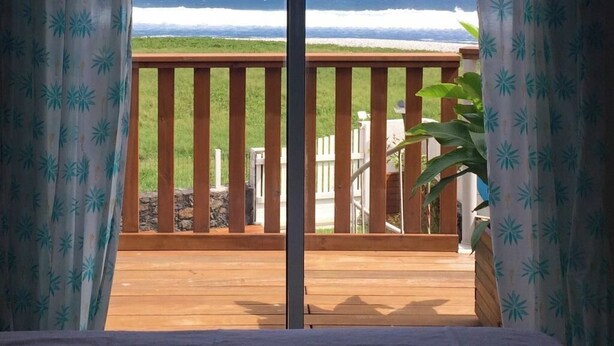 Blick durch eine Schiebetür auf ein Holzdeck mit Blick auf einen Strand und Wellen im Meer.