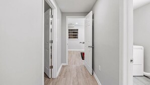 A narrow hallway with white walls and wooden flooring, leading to a room with a window and a door.